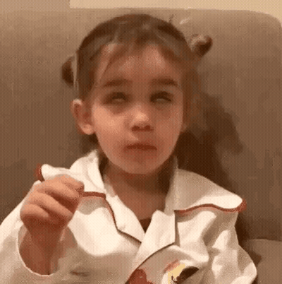 A Little Girl Is Sitting On A Couch With Her Eyes Closed And Eating A Cookie . GIF