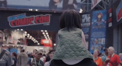 A Little Girl Is Sitting On A Person 'S Shoulders In Front Of A Sign That Says Comic Con GIF