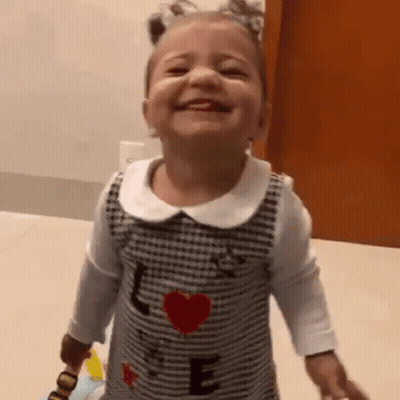 A Little Girl Is Smiling While Holding A Toy And Wearing A Dress With A Heart On It . GIF