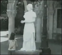 A Little Girl Is Standing In Front Of A Statue Of A Woman In A White Dress . GIF