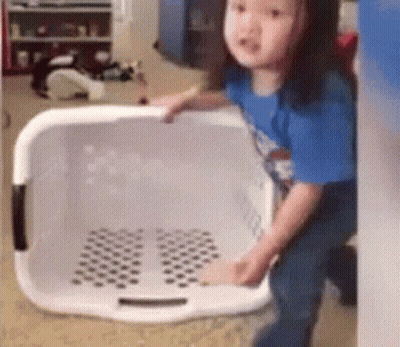 A Little Girl Is Standing Next To A Laundry Basket In A Room . GIF