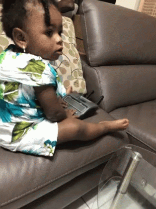 A Little Girl Sits On A Couch With A Remote Control In Her Hand GIF