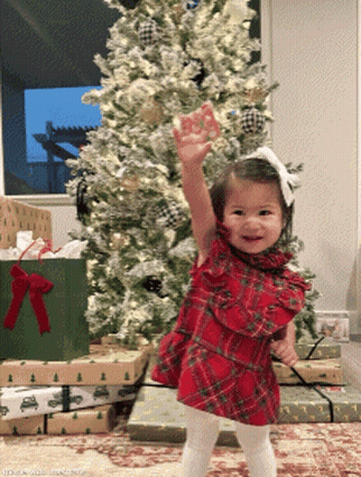 A Little Girl Stands In Front Of A Christmas Tree With Her Hand In The Air GIF