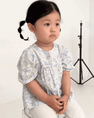 A Little Girl Wearing A Blue And White Floral Shirt Sits On A Clear Stool GIF