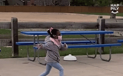 A Little Girl Wearing A Face Mask Is Walking In Front Of A Picnic Table . GIF