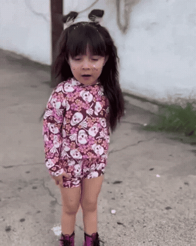 A Little Girl Wearing A Floral Shirt With Skulls On It Is Standing On A Sidewalk GIF