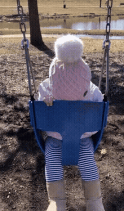 A Little Girl Wearing A Pink Hat And Striped Pants Is Sitting On A Blue Swing GIF