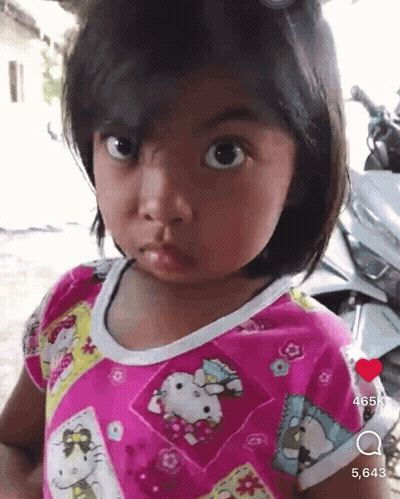 A Little Girl Wearing A Pink Hello Kitty Shirt Is Looking At The Camera GIF