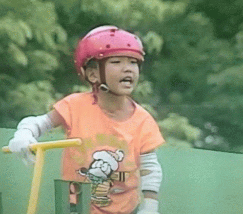 A Little Girl Wearing A Pink Helmet And An Orange Shirt With A Bear On It GIF