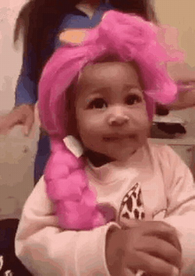 A Little Girl Wearing A Pink Wig And A Bow Is Sitting At A Table . GIF