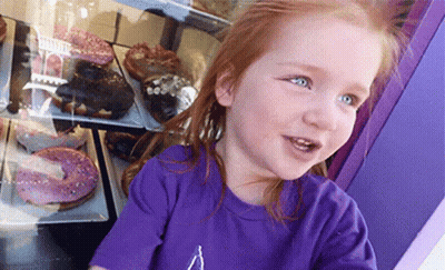 A Little Girl Wearing A Purple Shirt Is Looking At Donuts GIF