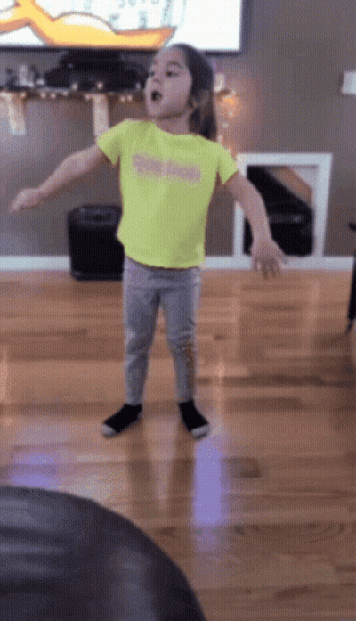 A Little Girl Wearing A Yellow Reebok Shirt Is Dancing In A Living Room GIF