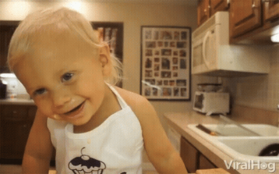 A Little Girl Wearing An Apron With A Cupcake On It Smiles In The Kitchen GIF
