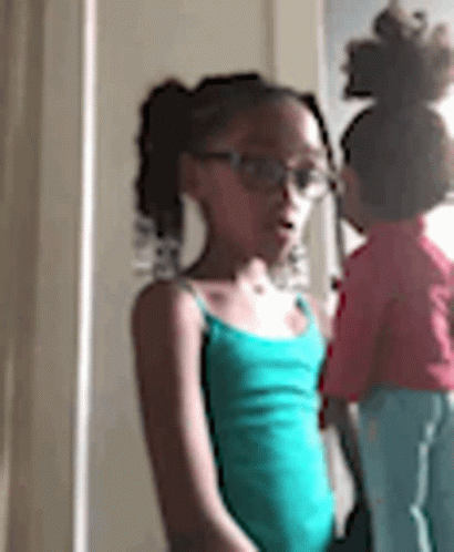 A Little Girl Wearing Glasses Is Standing Next To A Doll In A Room . GIF