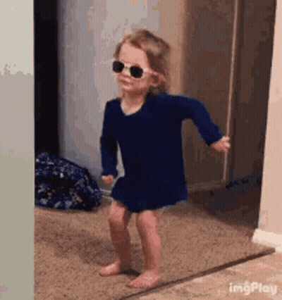 A Little Girl Wearing Sunglasses And A Blue Dress Is Dancing In A Hallway . GIF