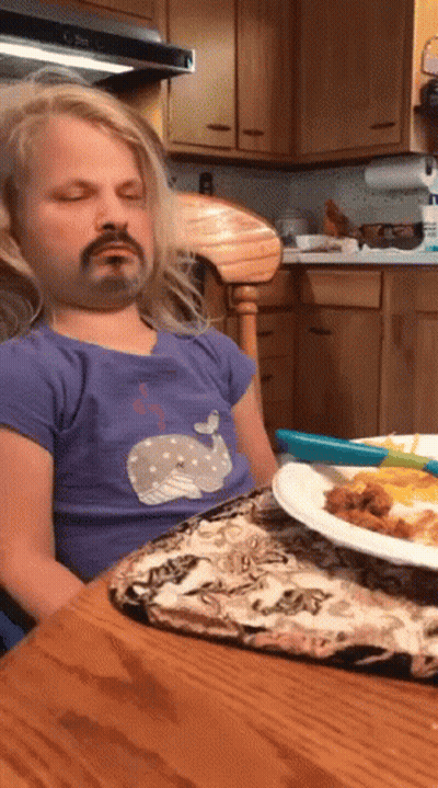 A Little Girl With A Fake Mustache Is Sitting At A Table With A Plate Of Food In Front Of Her GIF