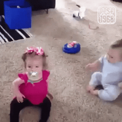 A Little Girl With A Pacifier In Her Mouth Is Sitting Next To A Baby . GIF