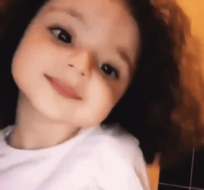 A Little Girl With Curly Hair Is Smiling And Looking Up At The Camera . GIF