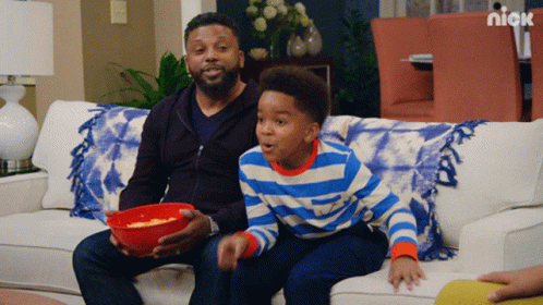 A Man And A Boy Are Sitting On A Couch With A Bowl Of Popcorn And A Nick Logo Behind Them GIF