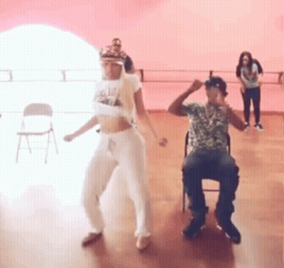 A Man And A Woman Are Dancing In A Dance Studio While A Man Sits In A Chair . GIF
