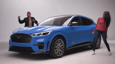 A Man And A Woman Are Dancing In Front Of A Blue Ford Mustang GIF