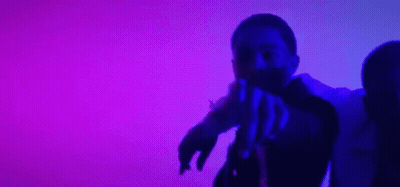 A Man And A Woman Are Dancing In Front Of A Purple Background . The Man Is Pointing At The Camera . GIF
