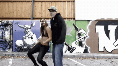 A Man And A Woman Are Dancing In Front Of A Wall With Graffiti Including The Letter N. GIF