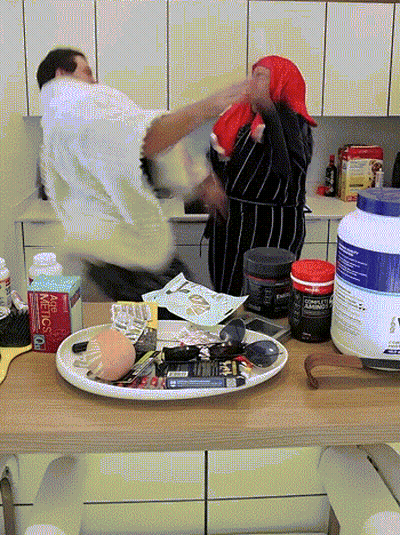 A Man And A Woman Are Fighting In A Kitchen With A Bottle Of Metcalf On The Counter Top GIF