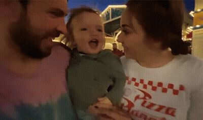 A Man And A Woman Are Holding A Baby . The Woman Is Wearing A Pizza Shirt . GIF