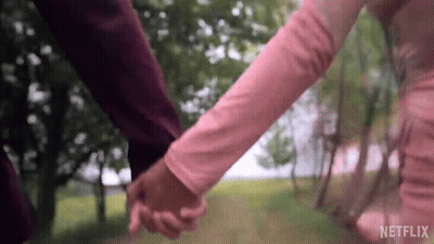 A Man And A Woman Are Holding Hands In A Park . The Woman Is Wearing A Pink Dress . GIF