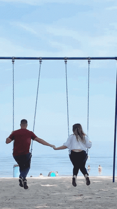 A Man And A Woman Are Holding Hands While Sitting On Swings GIF