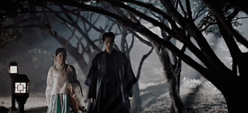 A Man And A Woman Are Holding Hands While Walking Through A Forest At Night . GIF