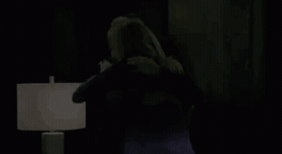 A Man And A Woman Are Hugging Each Other In A Living Room In Front Of A Lamp . GIF