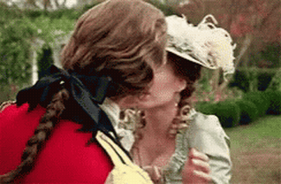 A Man And A Woman Are Kissing In A Garden . The Woman Is Wearing A Feathered Hat . GIF