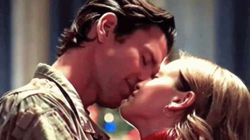 A Man And A Woman Are Kissing In A Room With A Christmas Tree In The Background . GIF