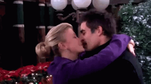 A Man And A Woman Are Kissing In Front Of A Christmas Tree . The Woman Is Wearing A Purple Jacket . GIF