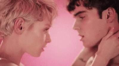 A Man And A Woman Are Kissing In Front Of A Pink Background . The Woman Is Touching The Man 'S Neck . GIF