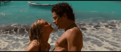 A Man And A Woman Are Kissing On A Beach With A Boat In The Background . GIF