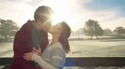 A Man And A Woman Are Kissing While Sitting On A Bench In Front Of A Field . GIF