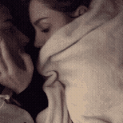 A Man And A Woman Are Kissing While Wrapped In A Blanket . GIF