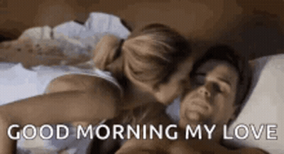 A Man And A Woman Are Laying In Bed And The Woman Is Kissing The Man On The Cheek . GIF