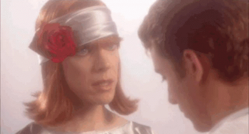 A Man And A Woman Are Looking At Each Other . The Woman Is Wearing A Headband With A Rose In Her Hair . GIF