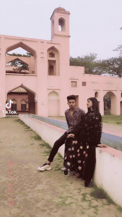 A Man And A Woman Are Posing For A Picture In Front Of A Pink Building With Tiktok Written On The Bottom GIF