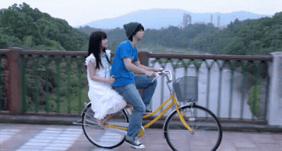 A Man And A Woman Are Riding A Bicycle On A Bridge Over A Body Of Water . GIF