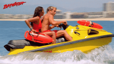 A Man And A Woman Are Riding A Yellow Jet Ski With Baywatch Written In Red GIF