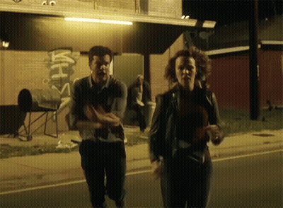 A Man And A Woman Are Running Down A Street In Front Of A Building That Has A Sign On It That Says ' With ' GIF