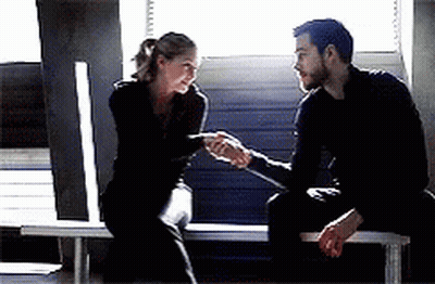 A Man And A Woman Are Shaking Hands While Sitting On A Bench In Front Of A Window . GIF