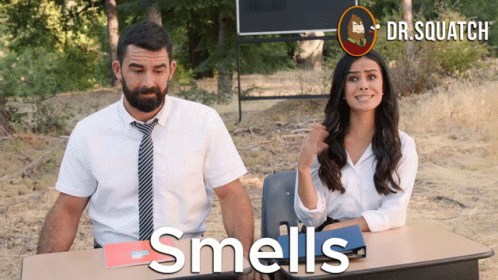 A Man And A Woman Are Sitting At A Desk With The Words Smells Written On The Table GIF