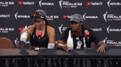 A Man And A Woman Are Sitting At A Table Drinking From Bottles GIF