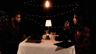 A Man And A Woman Are Sitting At A Table Drinking Wine And Talking GIF